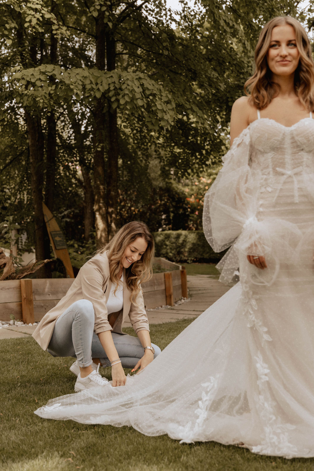 Michèle Tina Hug richtet die lange Schleppe des Brautkleides, während die Braut in einem Spitzenkleid im Garten steht.