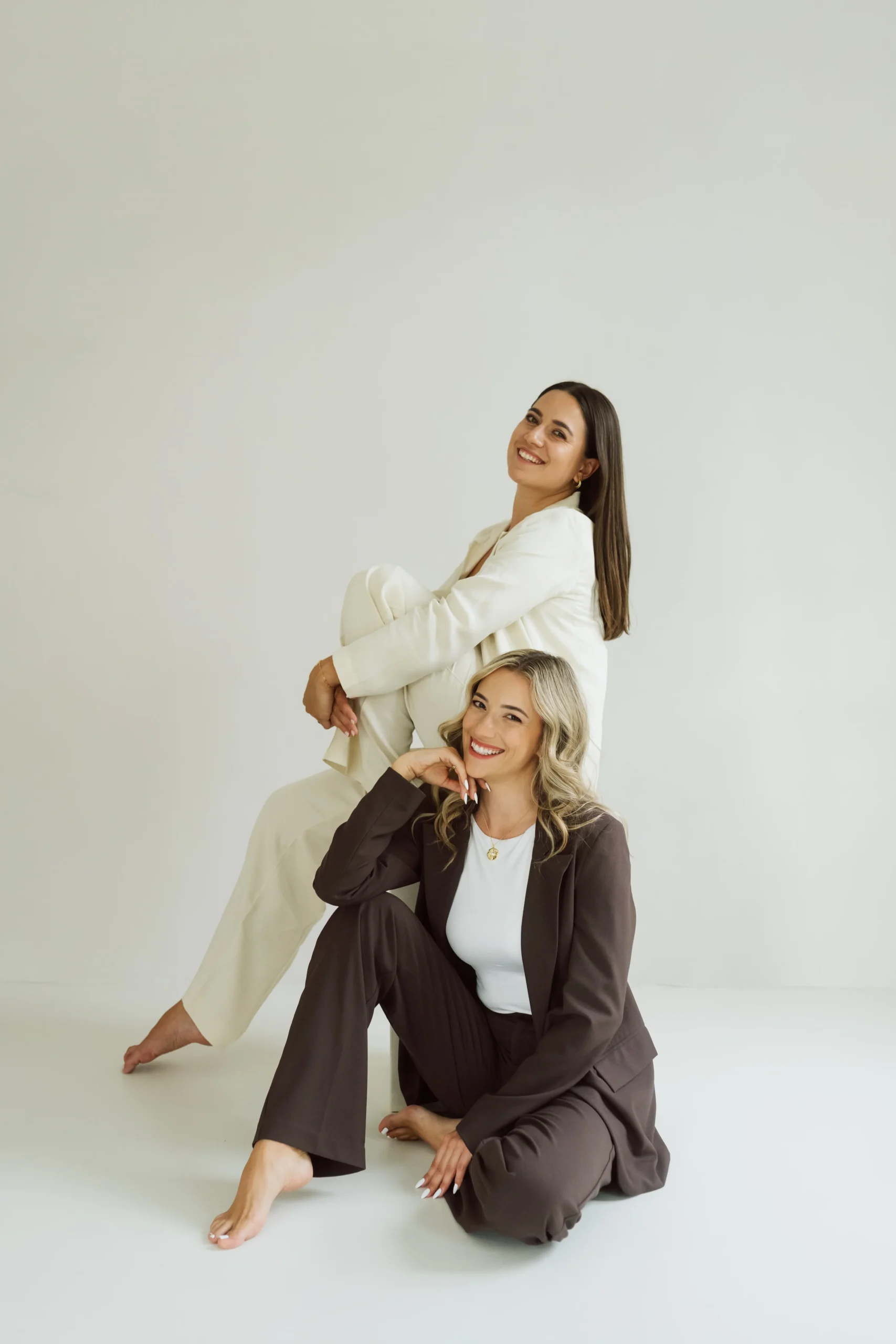 Michèle und Giuliana posieren gemeinsam in hellen Outfits vor einem neutralen Hintergrund.