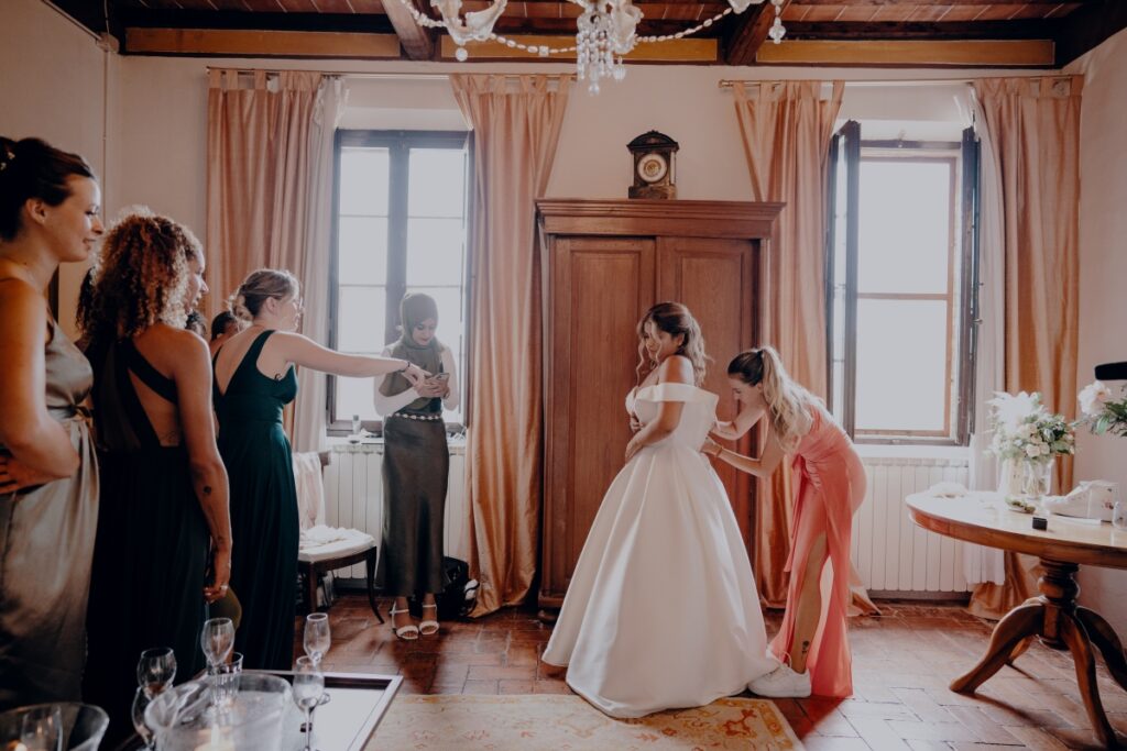 Braut in einem weissen Hochzeitskleid steht in einem hellen Zimmer, während mehrere Frauen um sie herum bei den Vorbereitungen helfen.