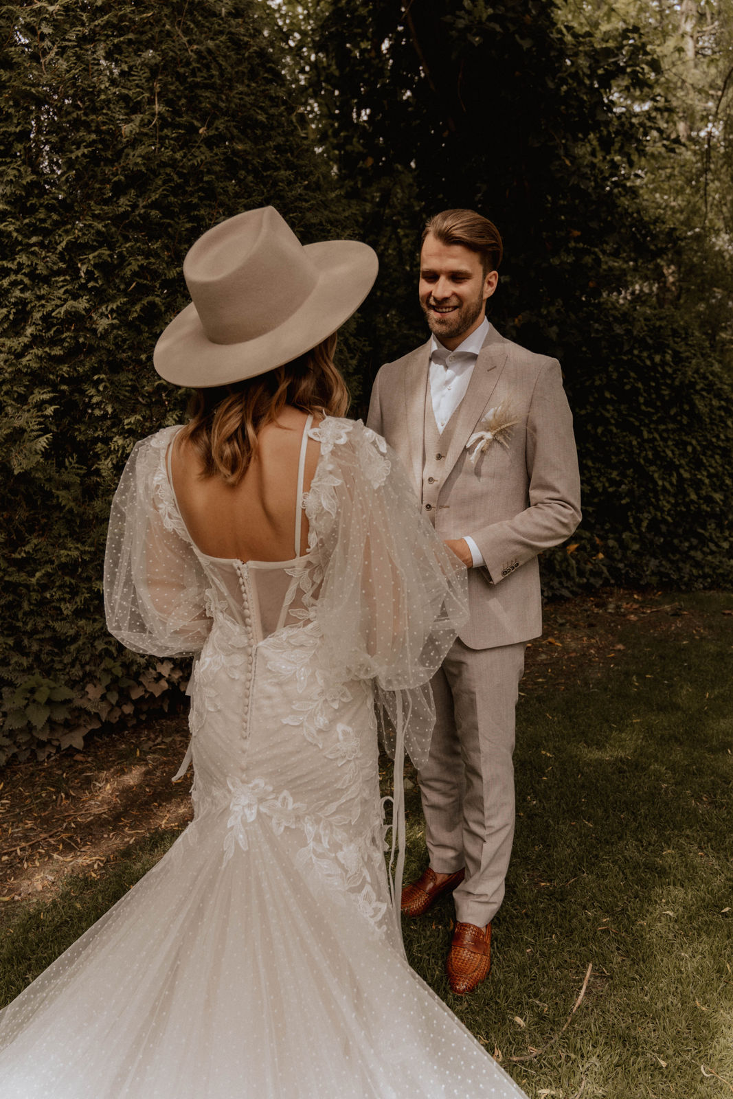 Braut mit beigefarbenem Hut und langem Spitzenkleid steht mit dem Bräutigam in einer natürlichen Gartenkulisse.