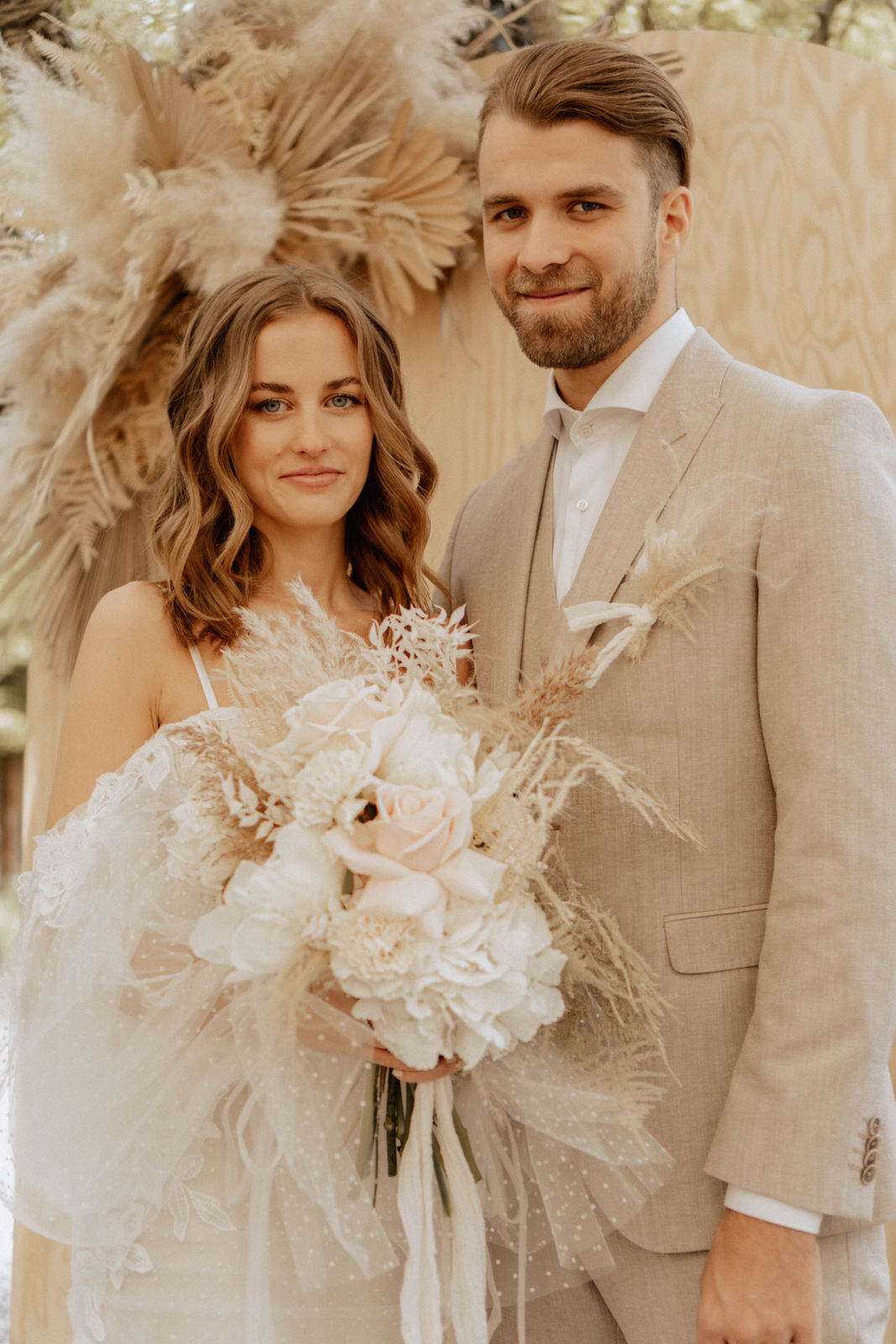 Boho-Brautpaar in heller Hochzeitskleidung mit einem üppigen Brautstrauss aus cremefarbenen Blumen und Pampasgras.