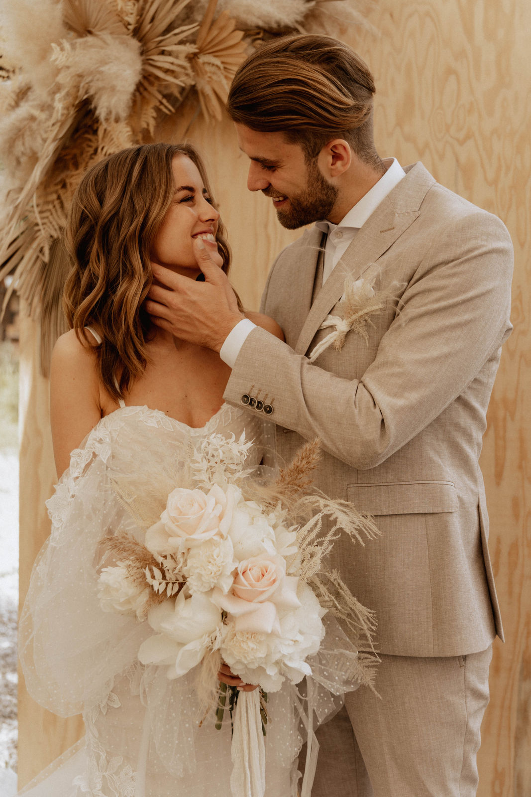Boho-Brautpaar in cremefarbener Hochzeitskleidung steht eng beieinander, während der Bräutigam die Wange der Braut berührt.