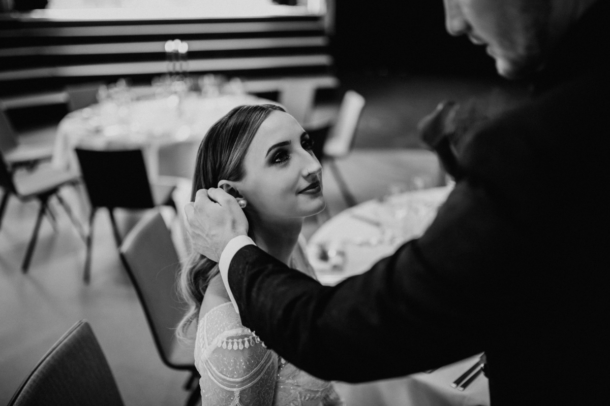 Schwarz-Weiss-Aufnahme einer lächelnden Braut, während ihr eine andere Person den Schmuck am Ohr richtet.