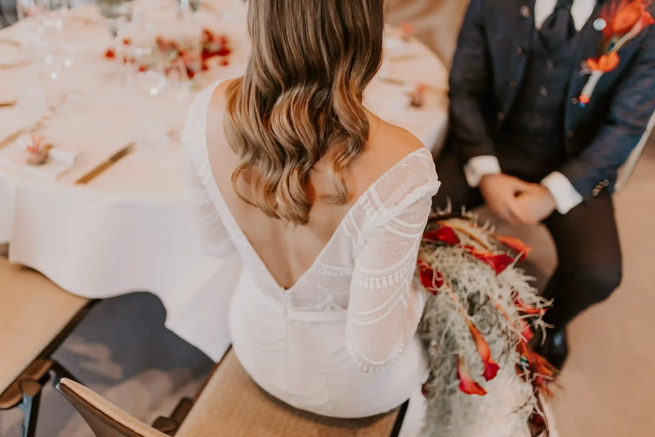Braut mit offenem, gewelltem Haar trägt ein weisses Kleid mit tiefem Rückenausschnitt und transparenten Ärmeln.