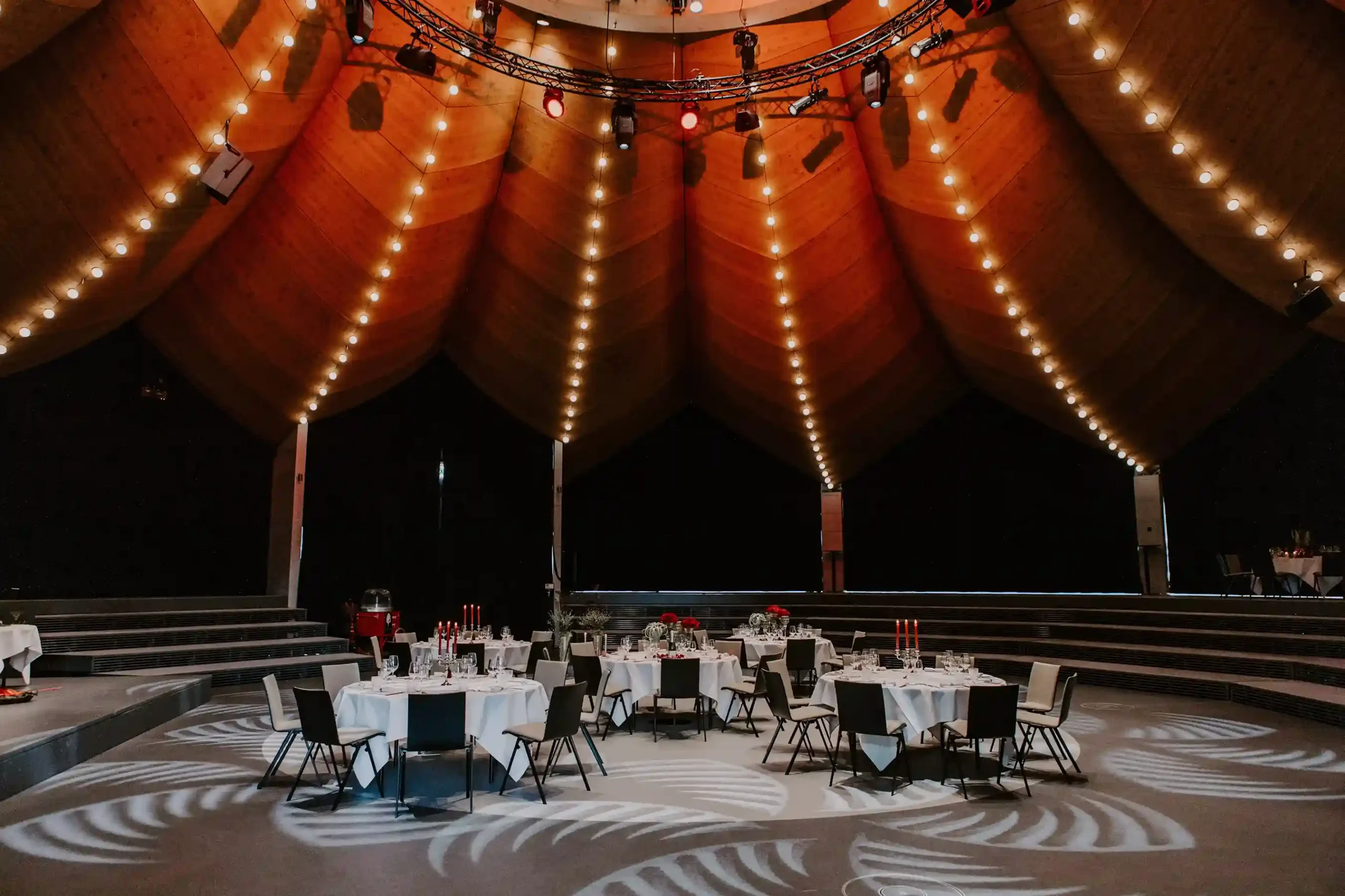 Festlich gedeckter Saal unter einem hohen rot-orangefarbenen Zeltdach mit langen Lichterketten und runden Hochzeitstischen.