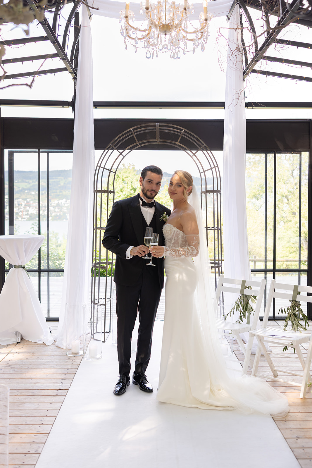 Sina und Damiano stehen mit Champagnergläsern unter einem filigranen Rundbogen in ihrer Hochzeitslocation.