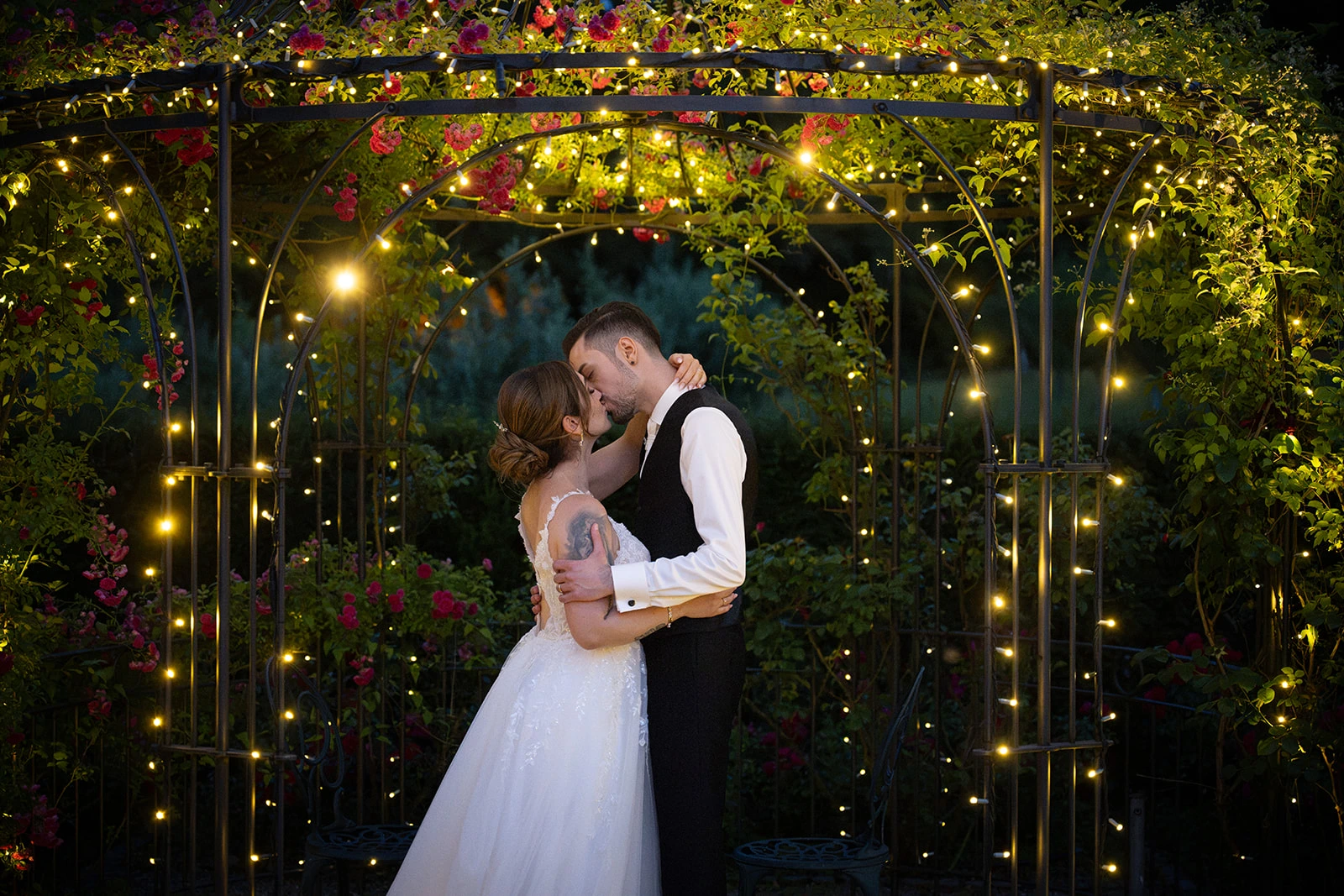 Valentina und Giacomo küssen sich unter einem mit Rosen und warmen Lichterketten geschmückten Gartenbogen.