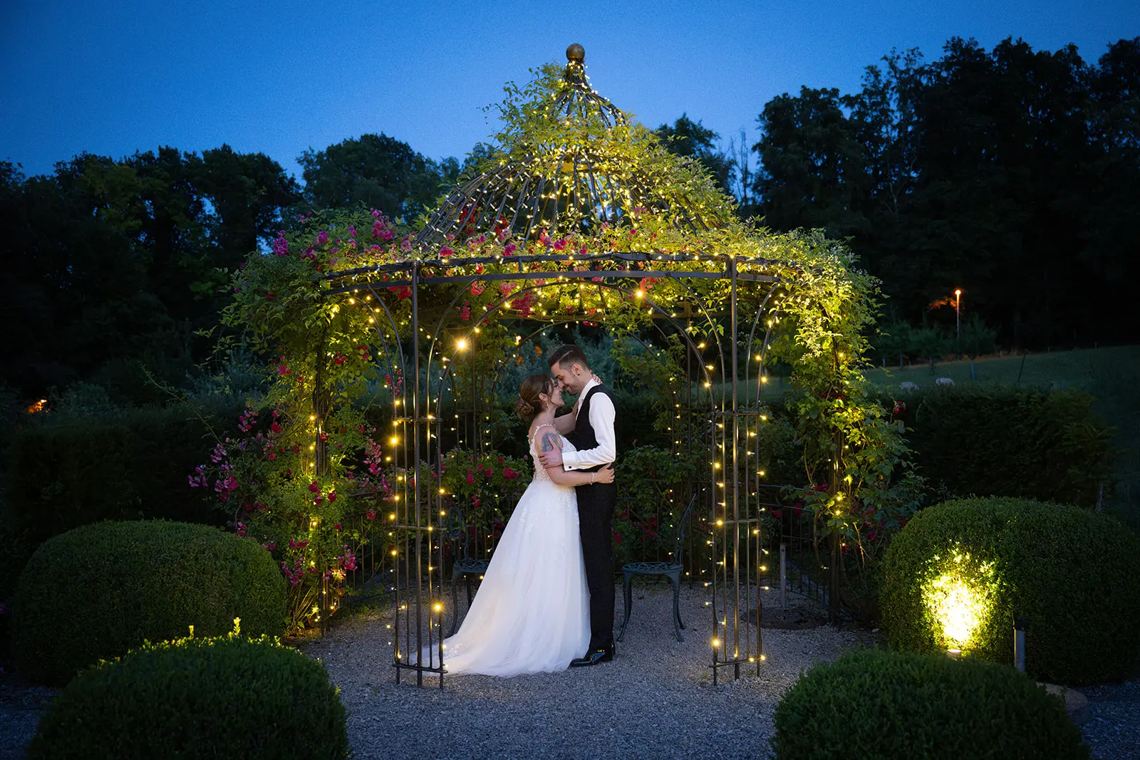 Valentina und Giacomo stehen eng umschlungen in einem mit Blumen und Lichterketten geschmückten Gartenpavillon.