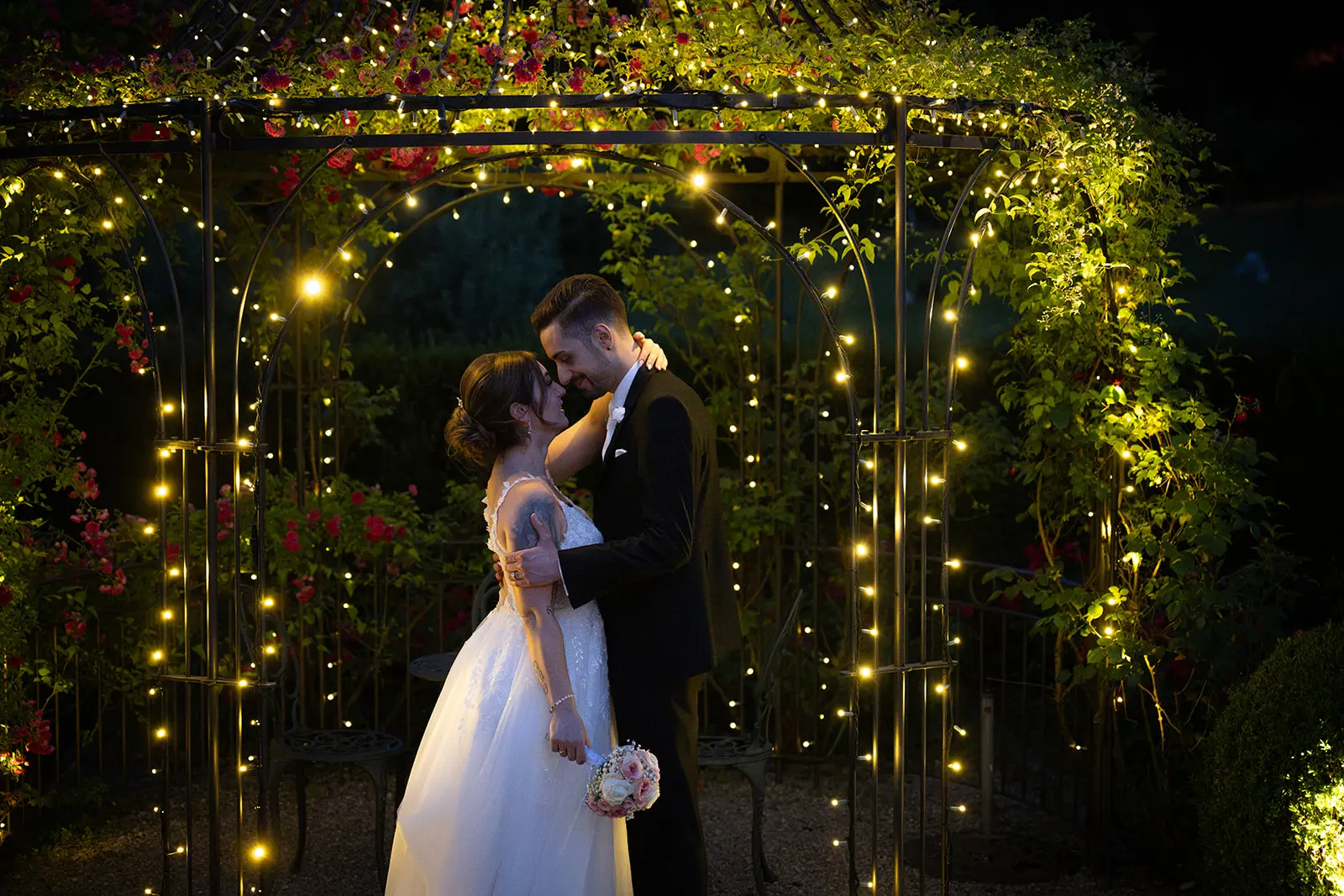 Valentina und Giacomo küssen sich vor einem mit warmen Lichterketten und Blumen geschmückten Gartenbogen.