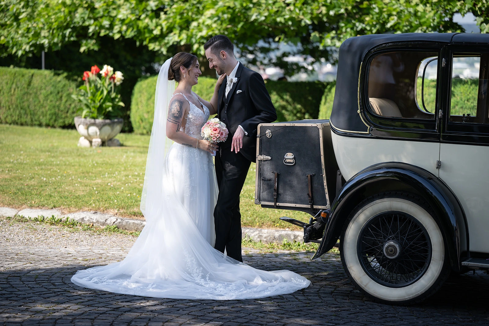 Valentina und Giacomo stehen lächelnd nebeneinander vor einem schwarzen Bentley Oldtimer im Grünen.