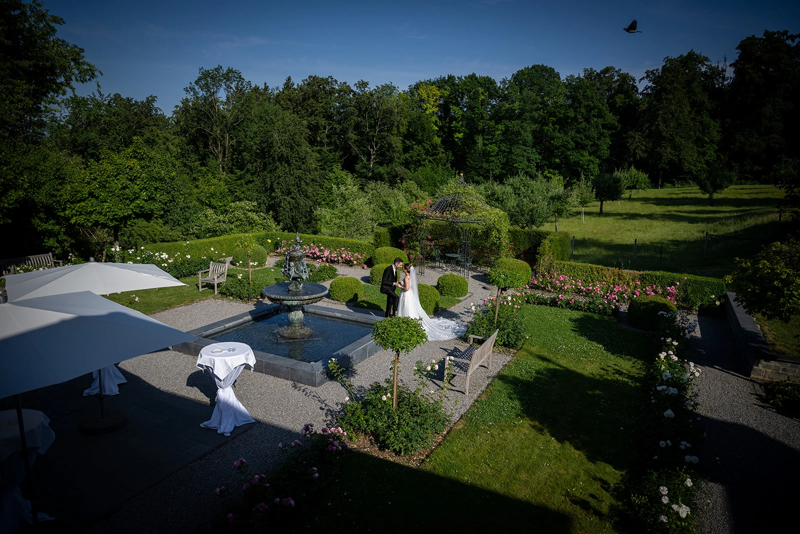 Blick von oben auf eine gepflegte Gartenanlage mit Brunnen, geschwungenen Wegen, Blumenbeeten und grünen Bäumen.