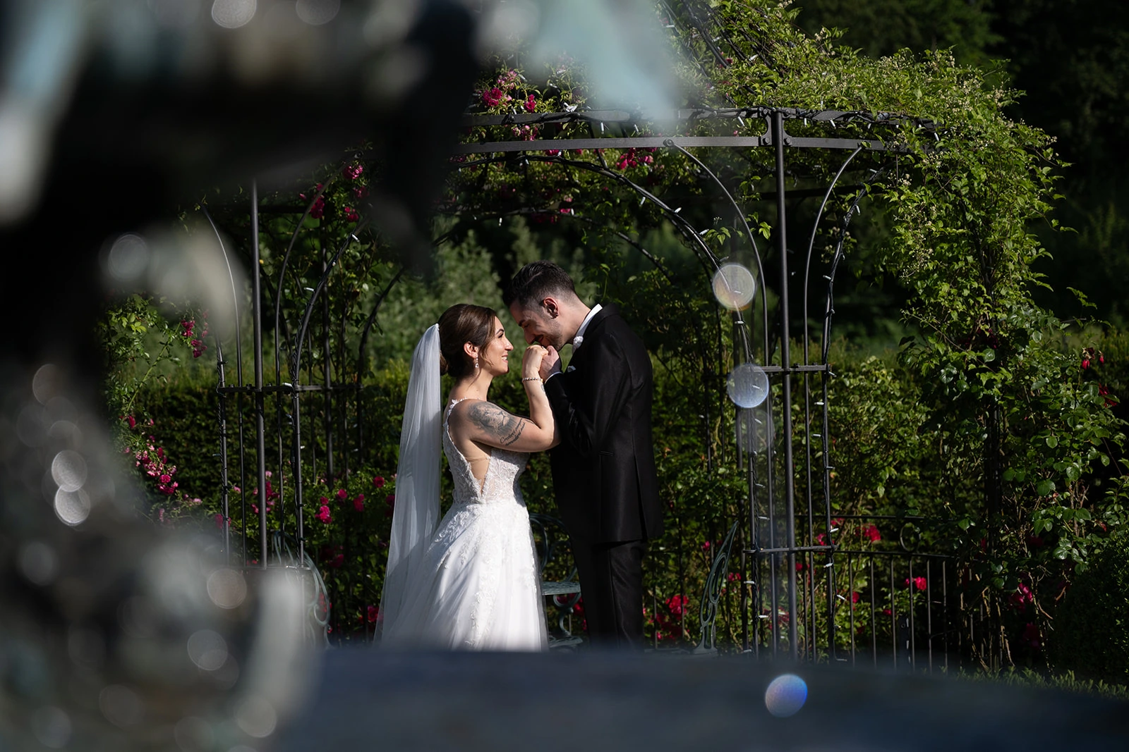 Valentina und Giacomo küssen sich in einem Garten vor einem mit Blumen geschmückten Torbogen.