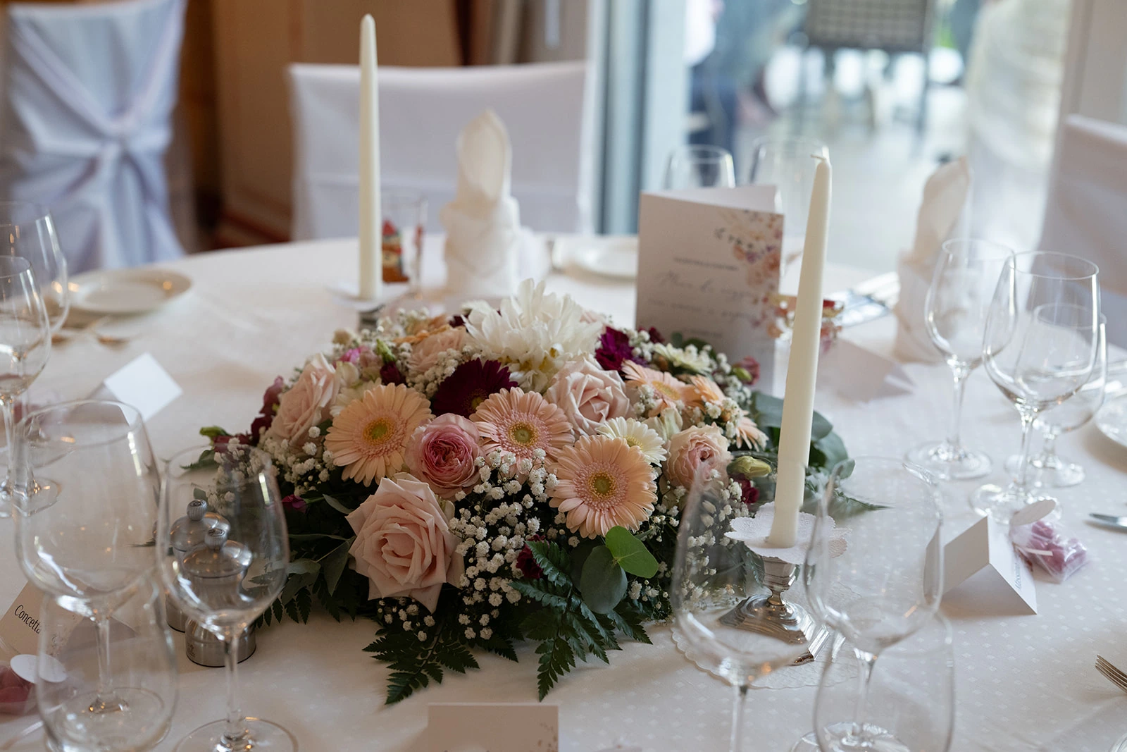 Üppiges Blumengesteck aus rosafarbenen Rosen, Gerbera und Schleierkraut auf einem festlich gedeckten Hochzeitstisch.