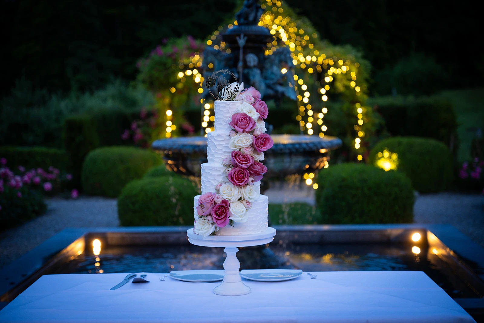 Mehrstöckige weisse Hochzeitstorte mit rosa und cremefarbenen Zuckerrosen vor einem beleuchteten Gartenbrunnen.
