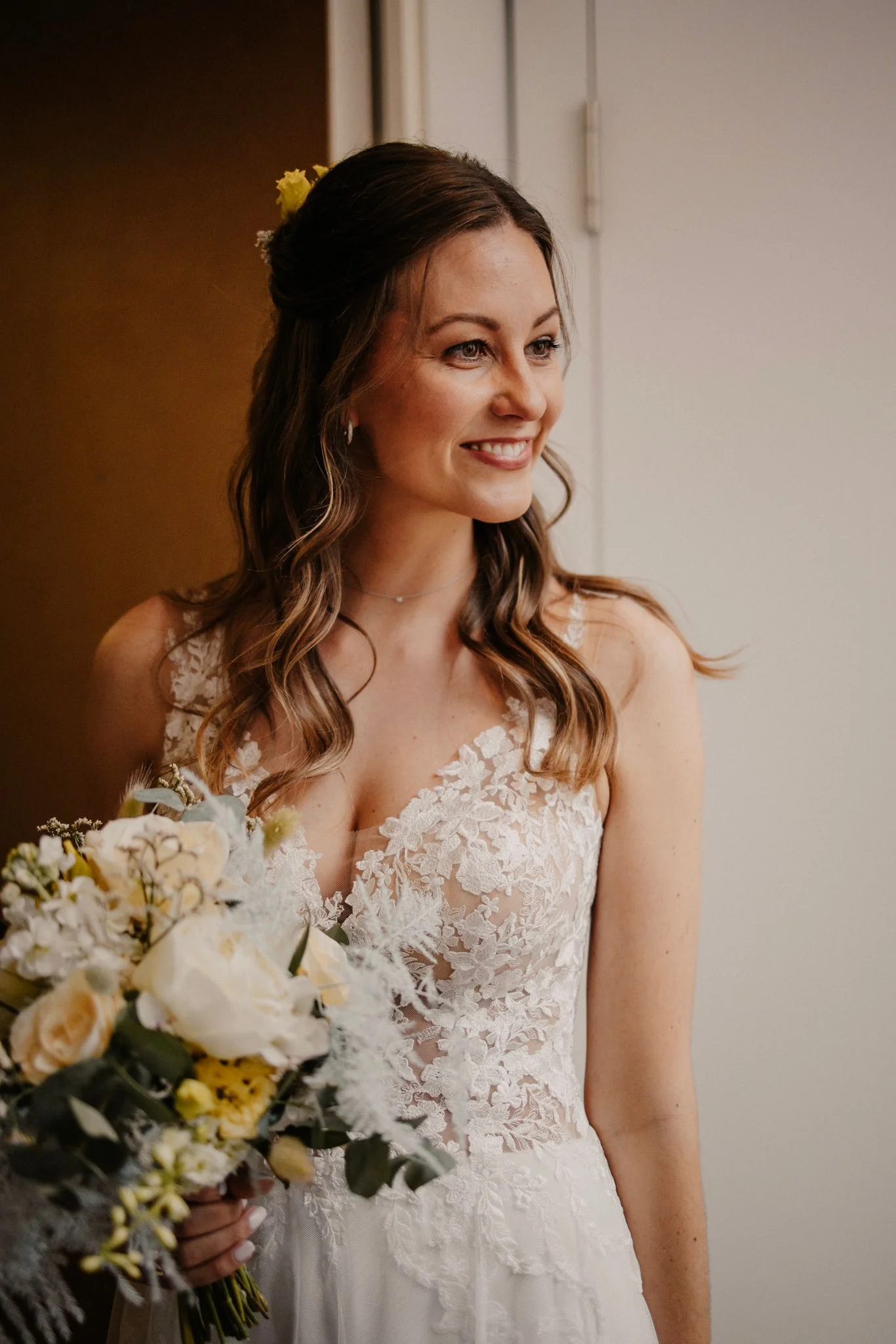 Céline lächelt in ihrem Brautkleid mit floraler Spitze und hält einen Strauss aus weissen und gelben Blumen.