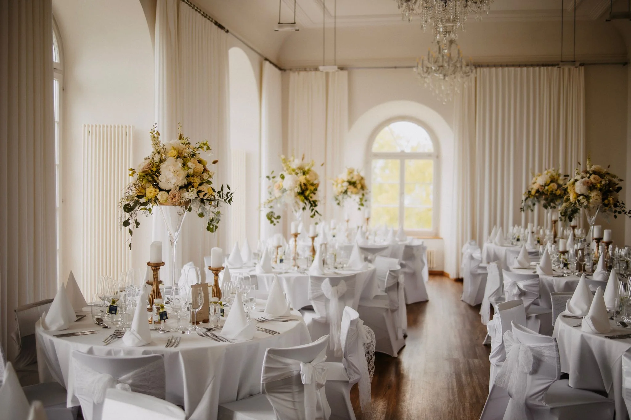 Eleganter Hochzeitssaal mit weiss gedeckten Tischen, Blumengestecken und hohen Rundbogenfenstern.