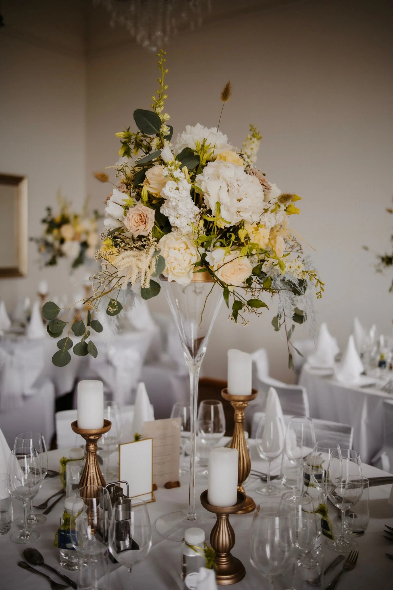 Üppiges Blumengesteck mit weissen und gelben Blüten, Kerzen und eleganten Gläsern auf einem Hochzeitstisch.