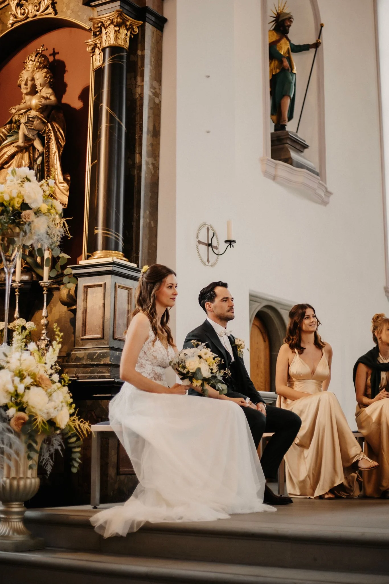 Céline und Philippe sitzen während ihrer kirchlichen Trauung nebeneinander, während weitere Personen neben ihnen Platz genommen haben.