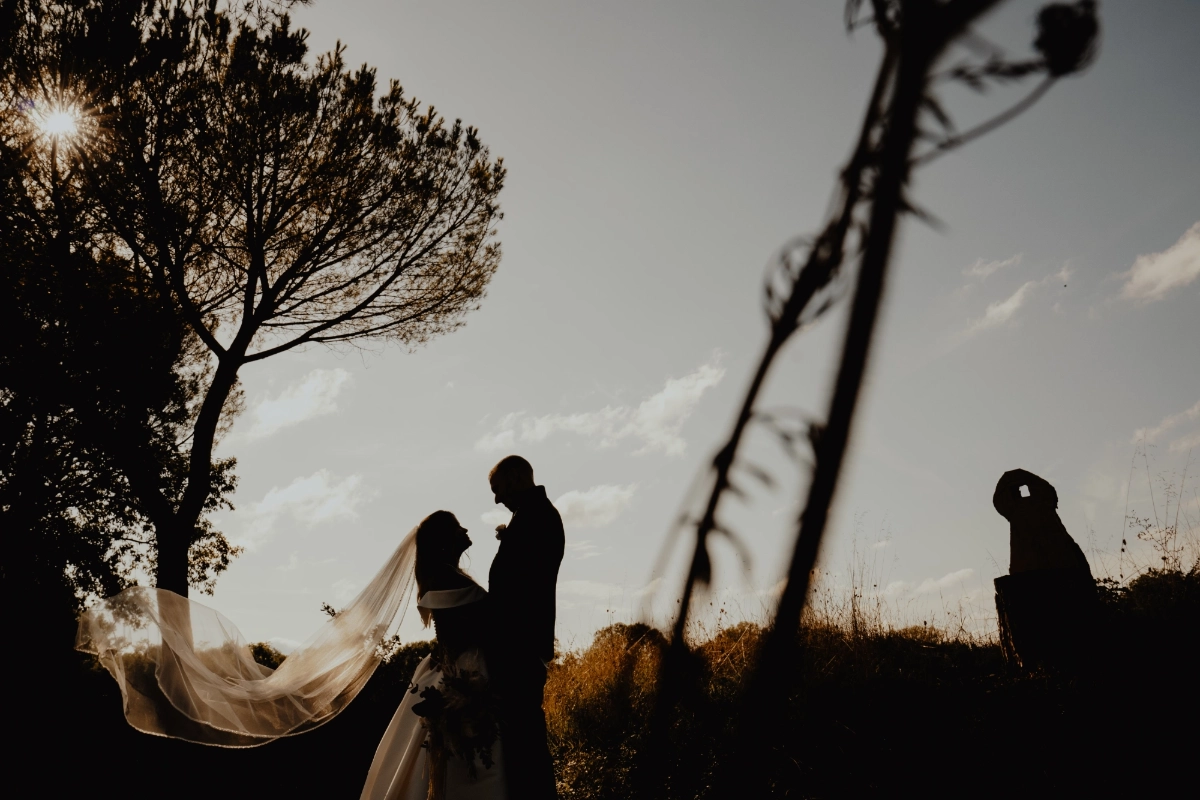 Silhouette von Thayna und Carson im Abendlicht zwischen Bäumen, während der Schleier der Braut im Wind weht.