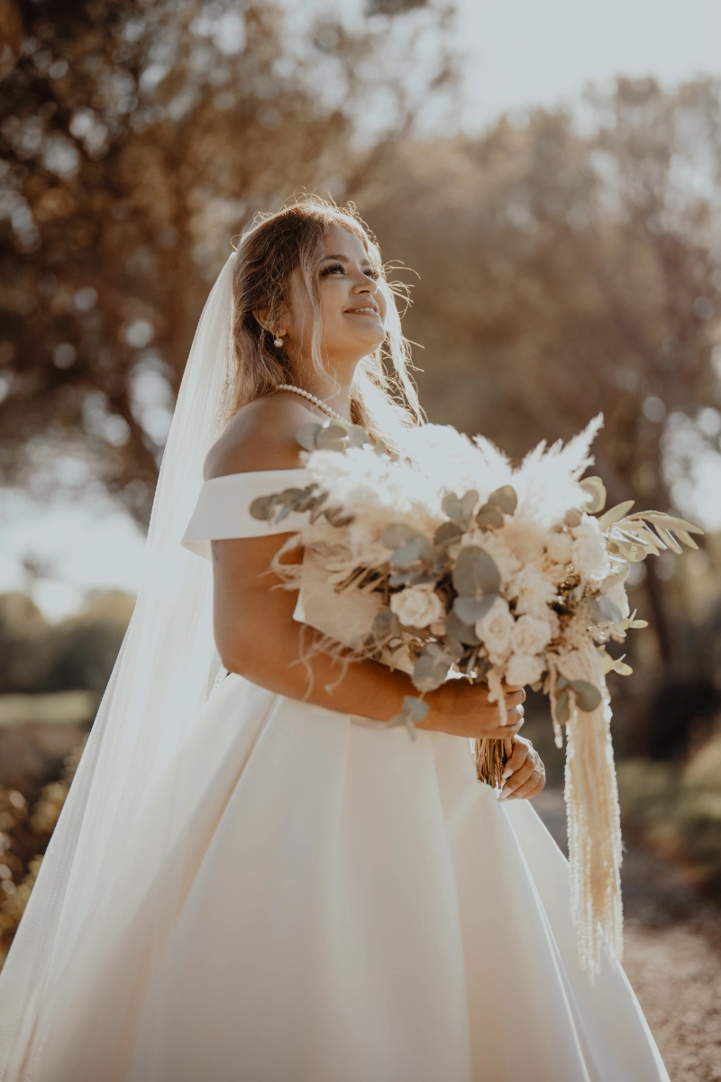 Thayna steht in ihrem weissen Brautkleid im warmen Sonnenlicht und hält einen grossen Blumenstrauss.