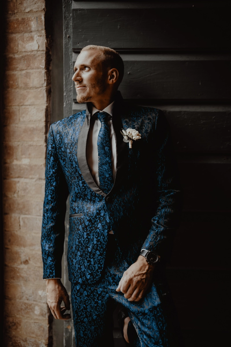 Carson steht in einem dunkelblau gemusterten Hochzeitsanzug mit Fliege und Ansteckblume im warmen Licht.