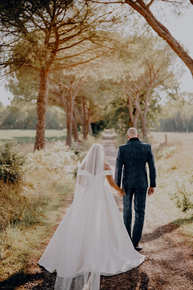 Thayna und Carson gehen Hand in Hand eine von Bäumen gesäumte Allee entlang, von hinten fotografiert.