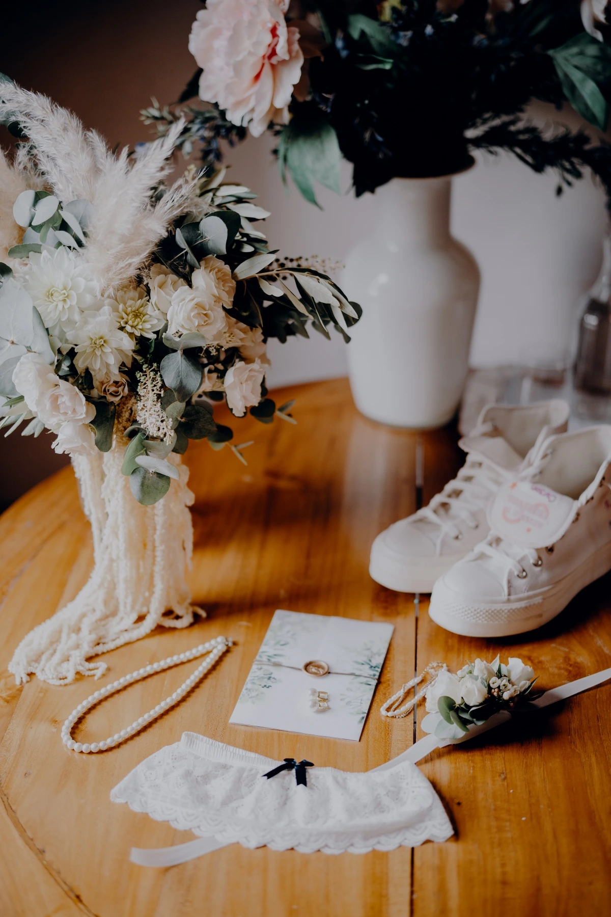 Hochzeitsdetails mit weissen Schuhen, Schmuck, Papeterie und einem Blumenstrauss auf einem Holztisch.